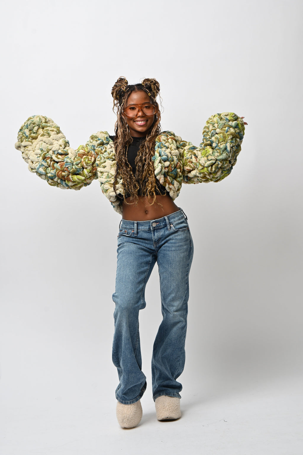 A model wearing a chunky, oversized cropped jacket made of textured, marled fabric in green, beige, and white, paired with blue jeans and beige slip-on shoes, posing against a white studio background.