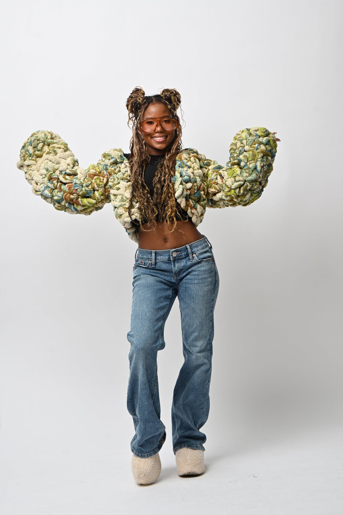 A model wearing a chunky, oversized cropped jacket made of textured, marled fabric in green, beige, and white, paired with blue jeans and beige slip-on shoes, posing against a white studio background.